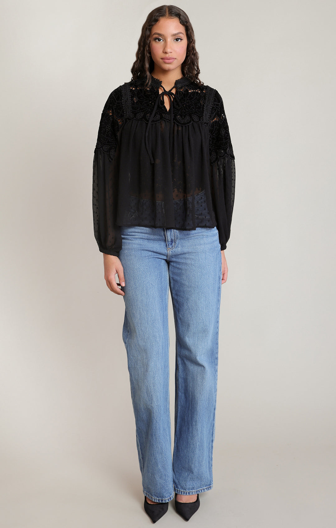 A woman modeling a black long-sleeve blouse with a lace-up keyhole neckline and sheer embroidered sleeves, paired with light-wash blue jeans and black pointed shoes, against a plain light gray studio background.