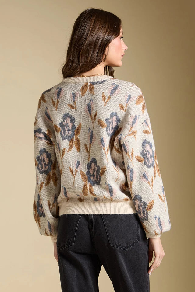 A woman shown from the back wearing a cream knit sweater with a blue and brown floral pattern, paired with black jeans, against a solid beige background.
