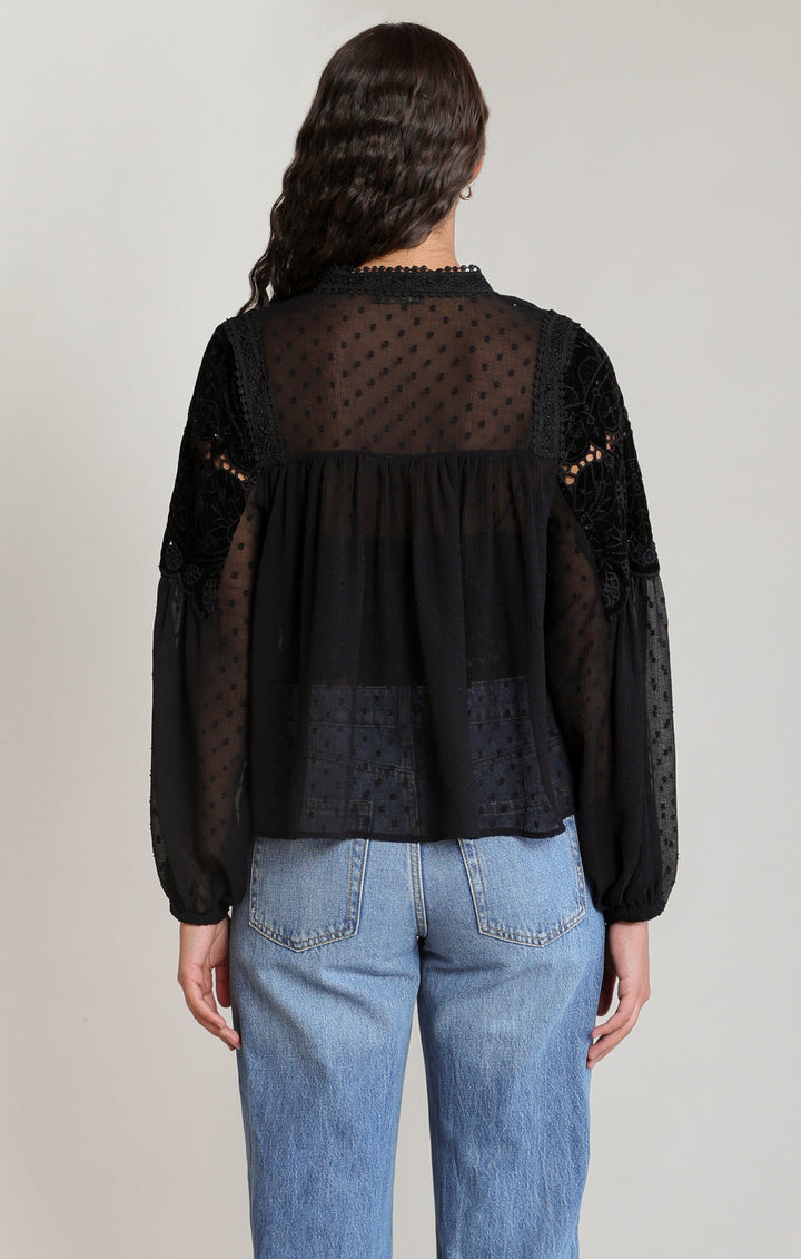 A woman viewed from the back wearing a black sheer long-sleeve blouse with a gathered ruffle waist and blue jeans, against a plain light gray studio background.