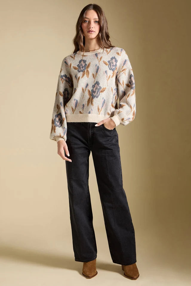 A woman modeling a cream-colored knit sweater with a blue and brown floral pattern, paired with black wide-leg jeans and brown suede boots, against a solid beige studio background.