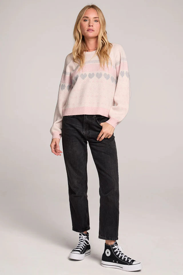 Woman wearing a light pink sweater with a pattern, black pants, and black Converse shoes on a plain background