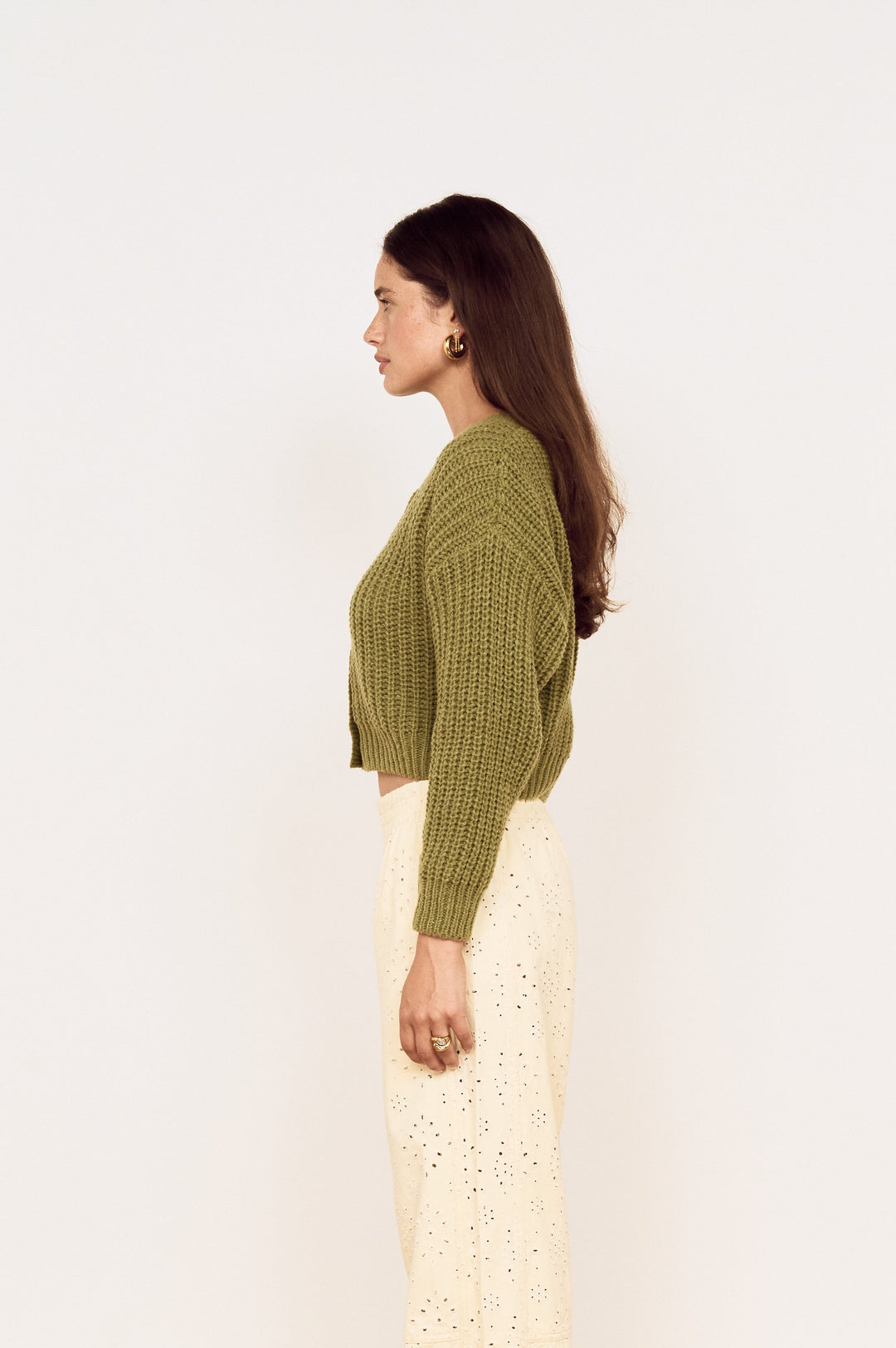 A woman stands in profile wearing a cropped olive green knit sweater and cream wide-leg lace-up pants on a light wood herringbone floor against a white wall.