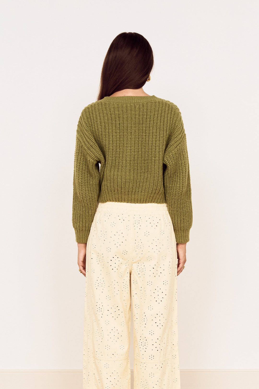 A woman viewed from behind wearing an olive green ribbed knit sweater and cream eyelet wide-leg trousers, standing on a light wood herringbone floor against a plain white wall.