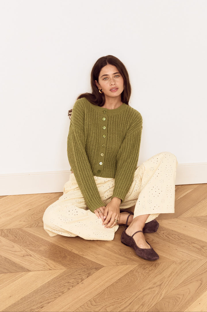 A woman sits cross-legged on a light wood herringbone floor against a white wall, wearing an olive green ribbed knit cardigan with buttons, cream wide-leg pants, and dark brown flats.