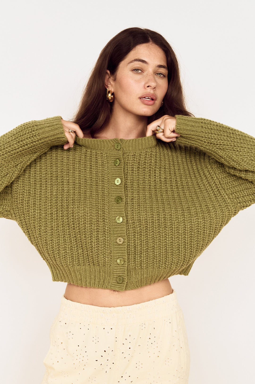 A woman wearing a cropped, olive green ribbed knit cardigan with wide batwing sleeves and a button-down front, paired with cream-colored eyelet pants, against a plain white studio background.