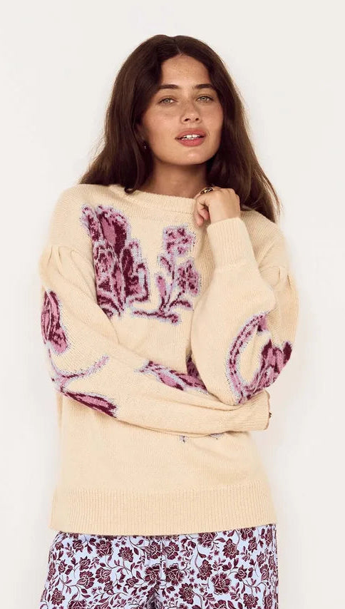 A woman modeling a cream-colored knit sweater with purple paisley patterns on the sleeves and chest, paired with matching patterned pants, against a plain white background.
