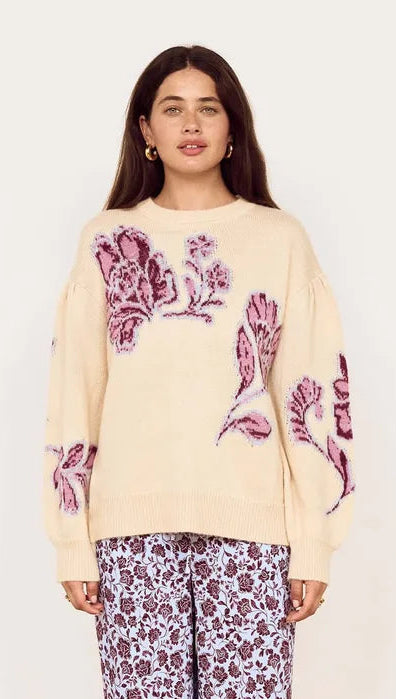 A woman stands in a room wearing a cream-colored knit sweater with purple floral embroidery and matching purple floral print wide-leg pants.