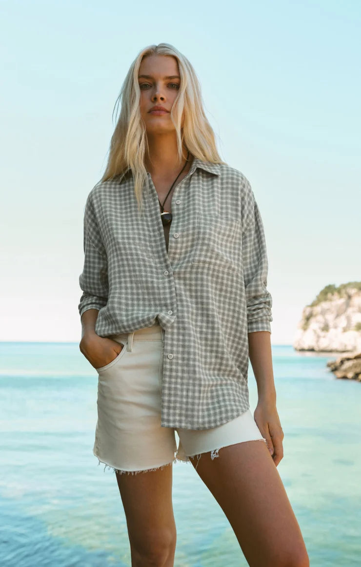 A woman standing on a beach wearing a light gray and white gingham button-up shirt and off-white denim shorts, with the ocean and rocky coastline in the background.
