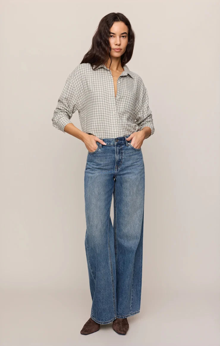 A woman stands against a plain light beige studio background wearing a light gray and white gingham button-up shirt, high-waisted wide-leg blue jeans, and dark brown pointed-toe flats.