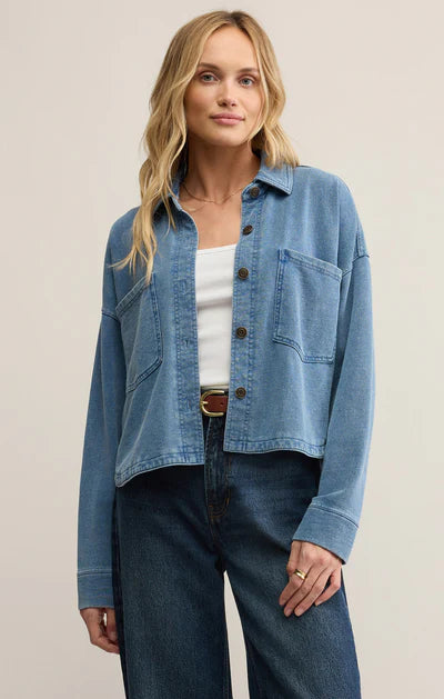Woman wearing a blue denim jacket over a white top and dark jeans on a beige background