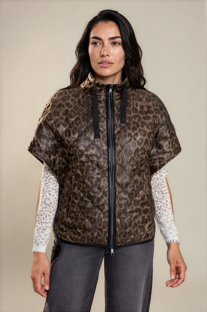 A woman modeling a brown and black leopard-print quilted hooded jacket with short sleeves, worn over a white patterned long-sleeve shirt and dark gray pants, against a solid beige studio background.