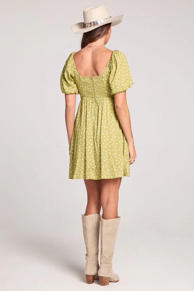 Woman wearing a green dress with puffed sleeves, beige boots, and a white hat on a light gray background