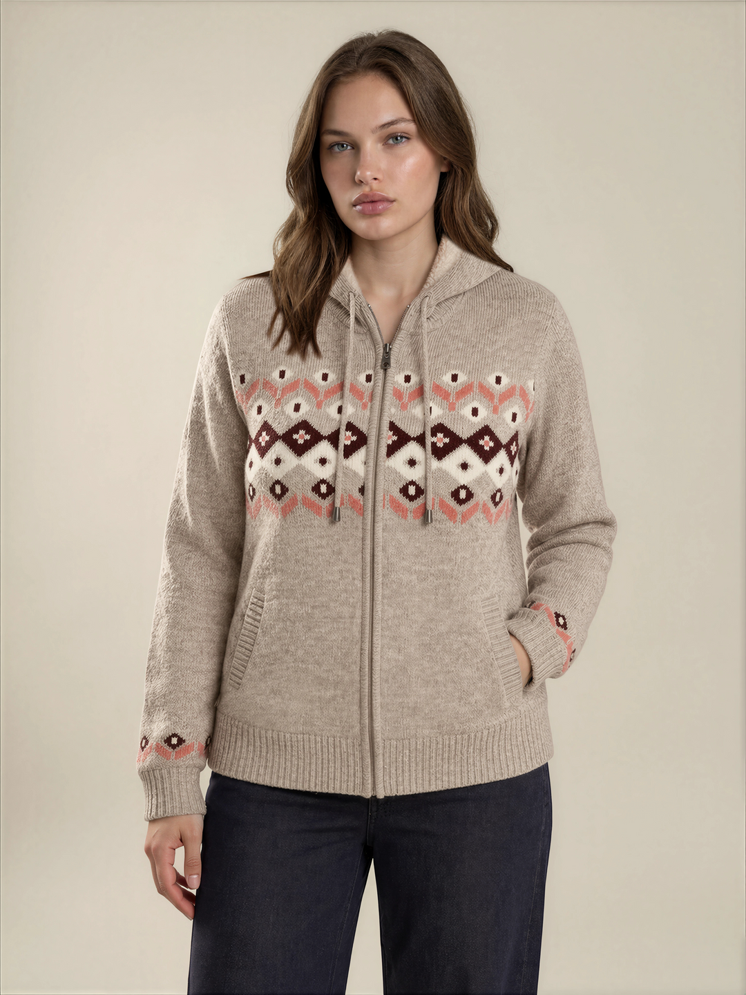 A woman wearing a beige zip-up hoodie with a colorful geometric pattern across the chest and cuffs, paired with dark blue pants, against a solid light beige background.