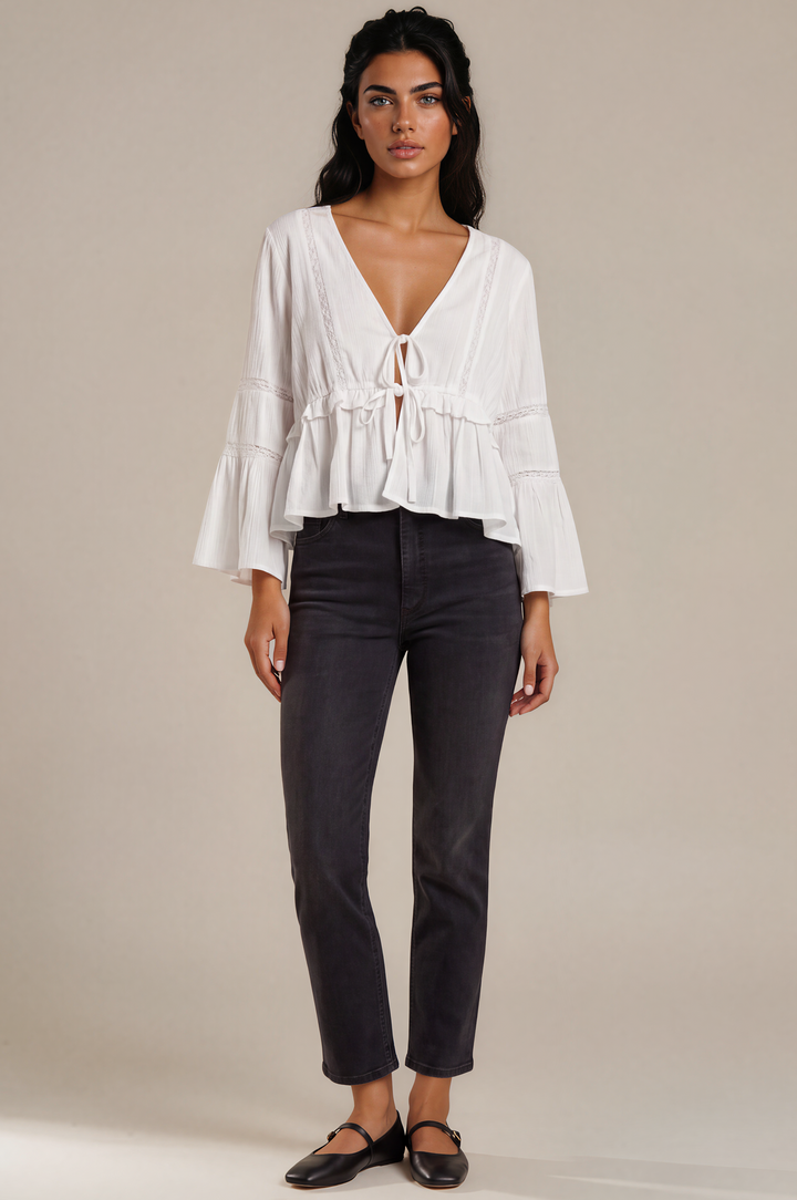 A woman modeling a white long-sleeve blouse with a deep V-neck, tie-front detail, and ruffled layers, paired with dark charcoal cropped pants and black flats, against a plain beige studio background.
