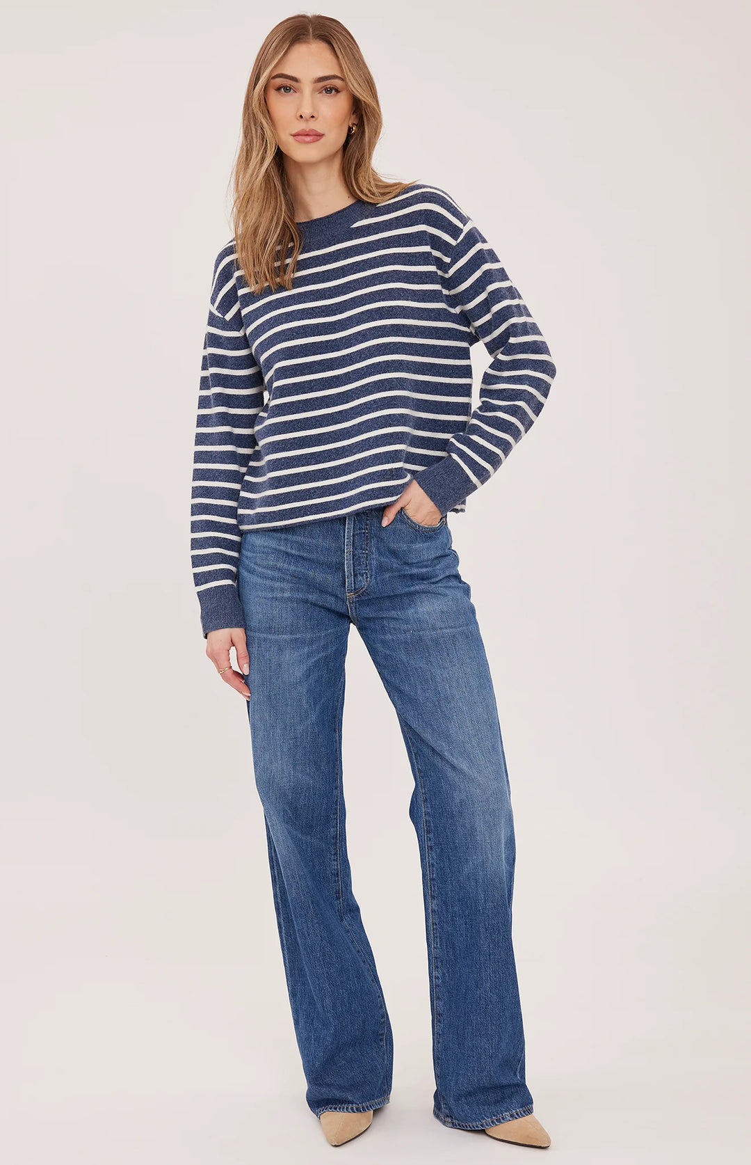 A woman modeling a navy and white horizontally striped long-sleeve knit sweater paired with blue jeans and beige pointed-toe flats, set against a plain light gray studio background.