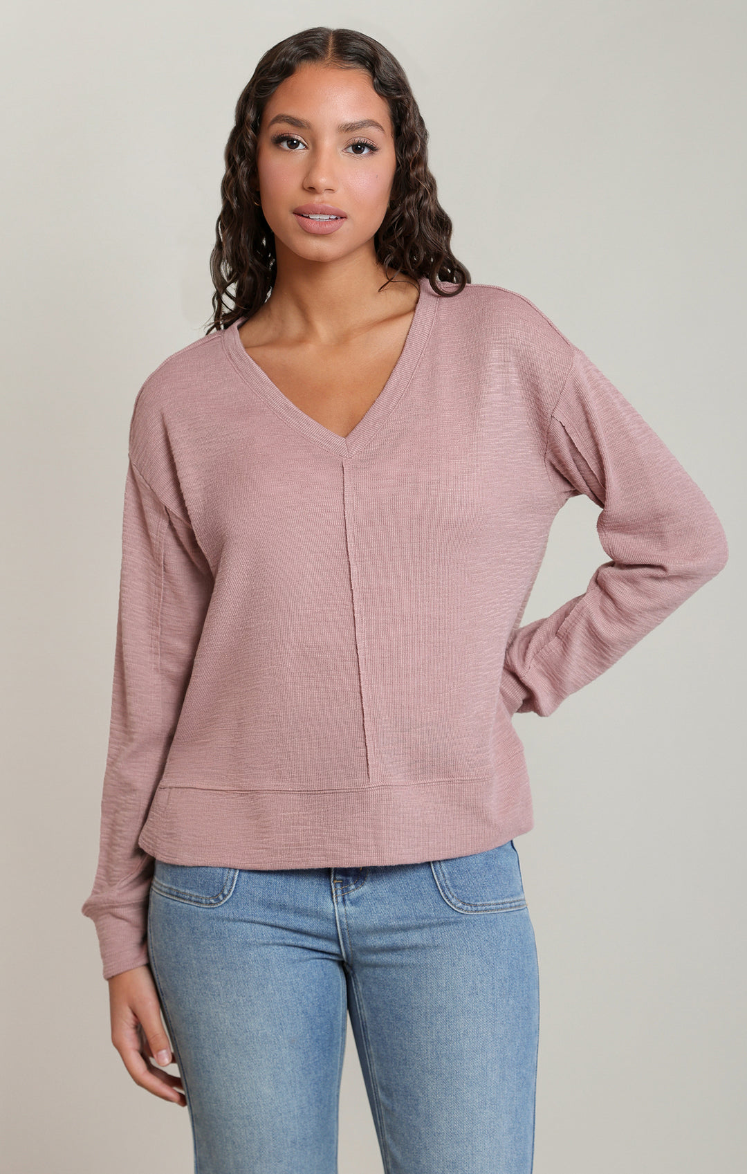 Woman wearing a mauve long-sleeve top and blue jeans on a plain background