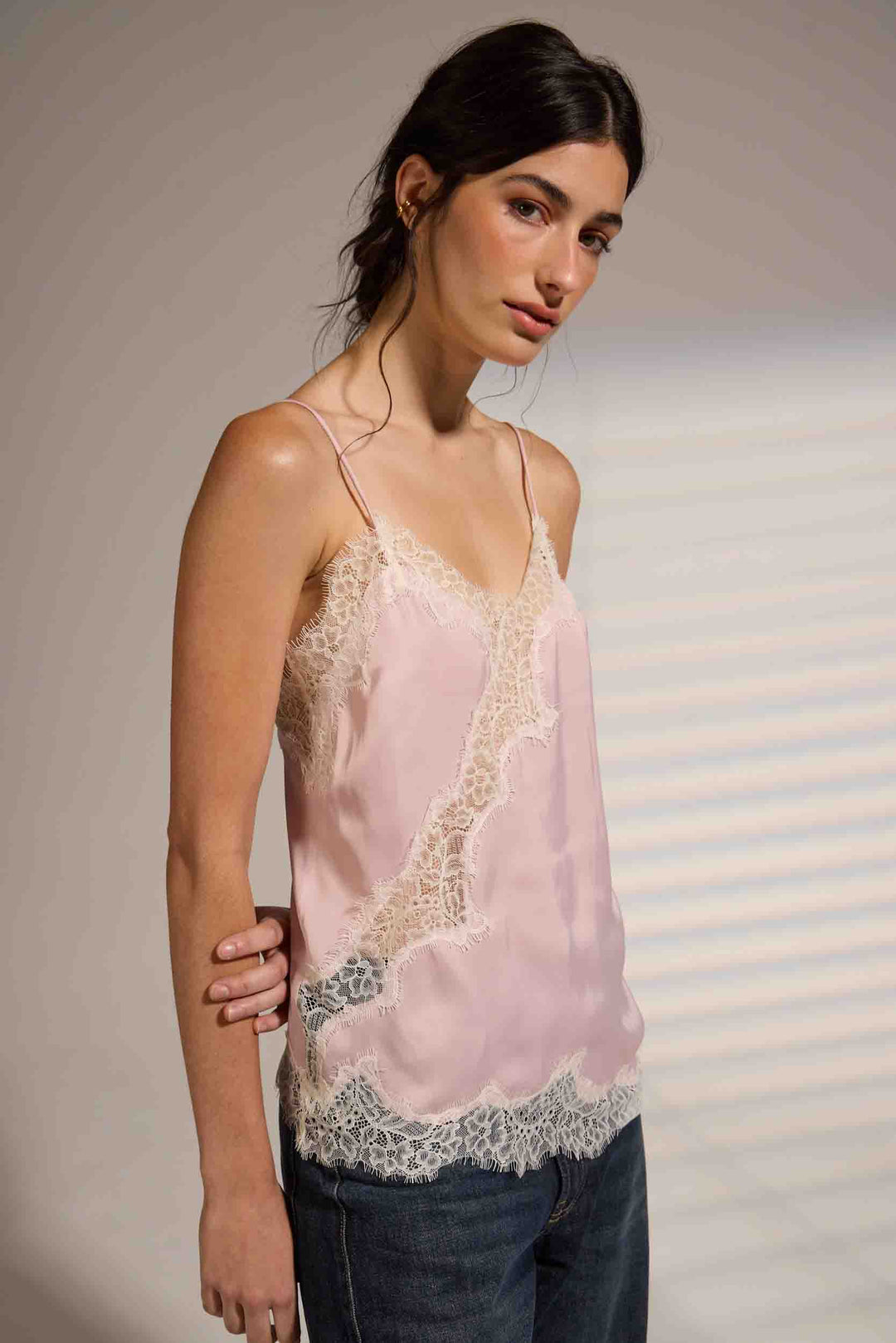 A woman modeling a light pink silky camisole top with white lace trim along the neckline, side panels, and hem, paired with dark blue jeans against a neutral studio backdrop.