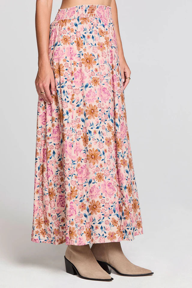 A model wearing a high-waisted, ankle-length maxi skirt with a pink, orange, and blue floral pattern, paired with beige ankle boots, against a light gray studio background.