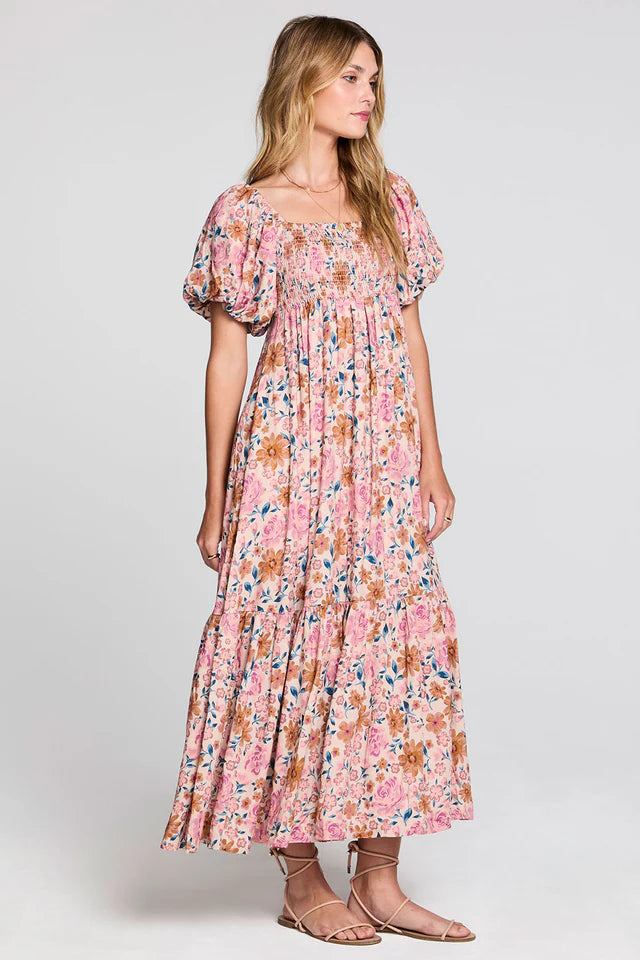 A woman modeling a pink floral maxi dress with puff sleeves and a smocked bodice, paired with tan flat sandals, against a light gray studio background.