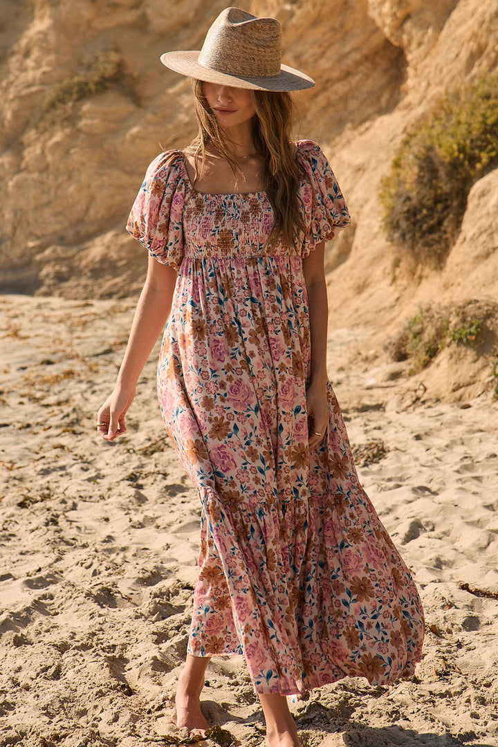 Woman in a floral dress and hat standing on sandy terrain with rocky background