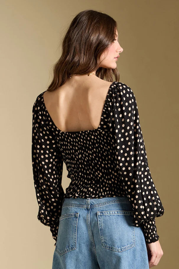 Woman wearing a black polka dot blouse with an open back and blue jeans on a beige background