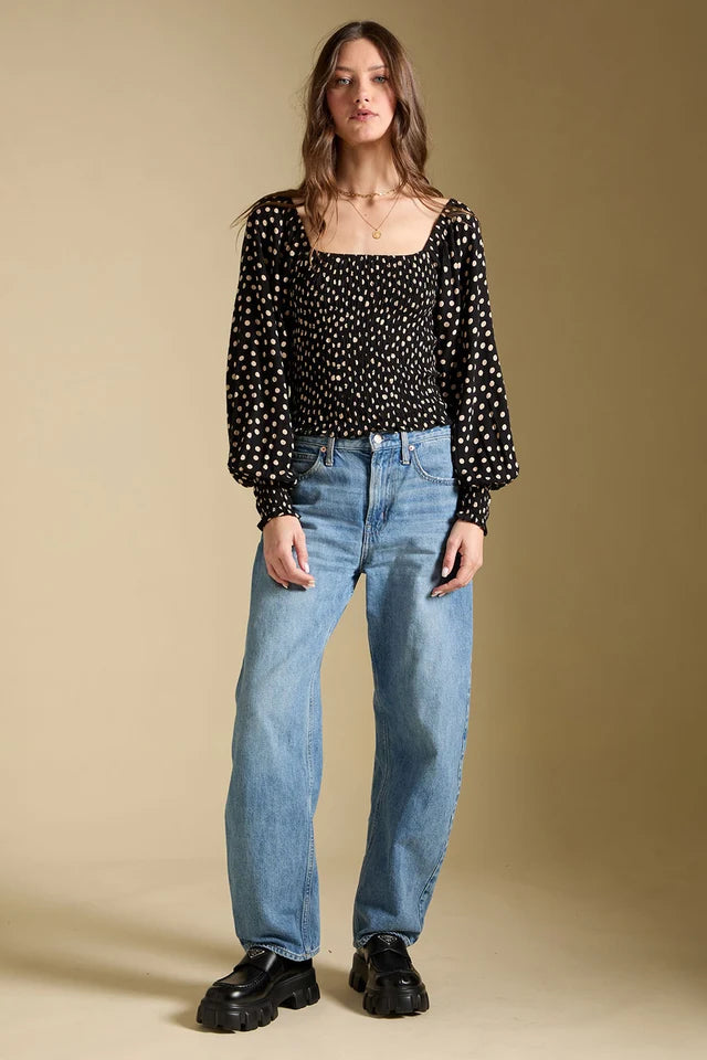 Woman wearing a black polka dot blouse and blue jeans against a beige background