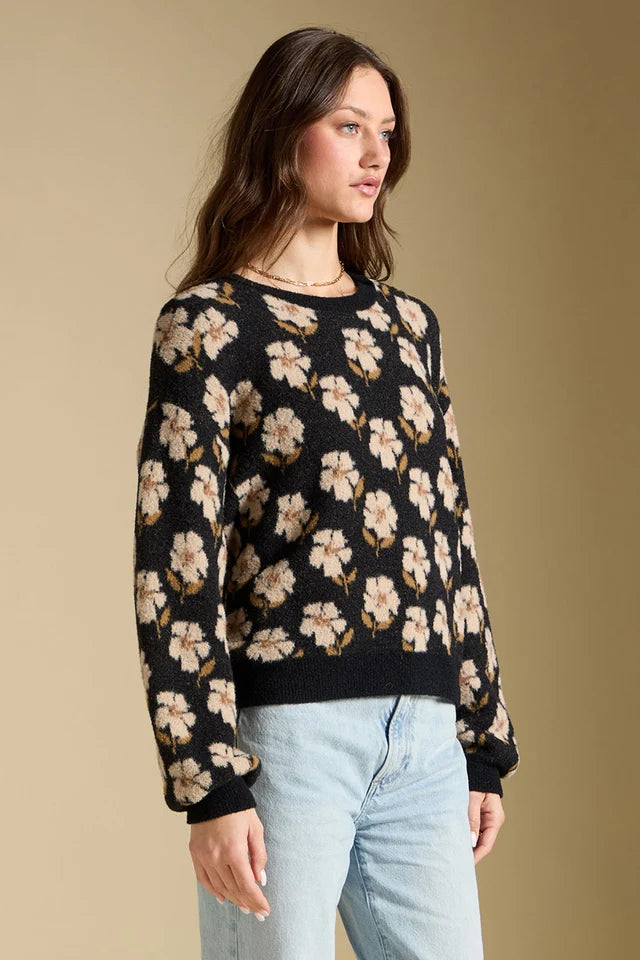 A woman wearing a black knit sweater with a beige and white floral pattern, paired with light blue jeans, standing against a solid beige background.