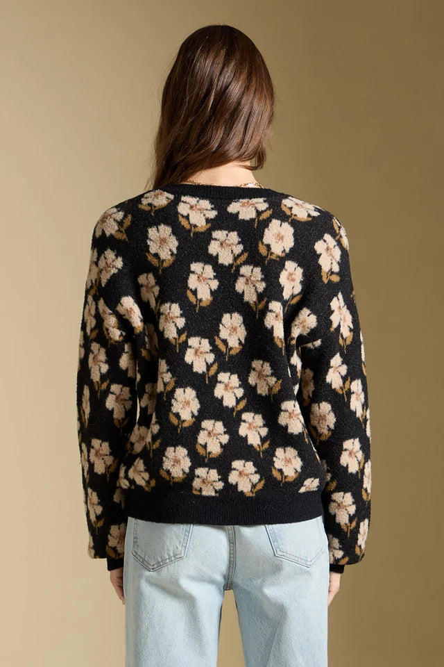 A woman viewed from the back wearing a black knit sweater with a beige and cream floral pattern, paired with light blue jeans, against a solid beige background.
