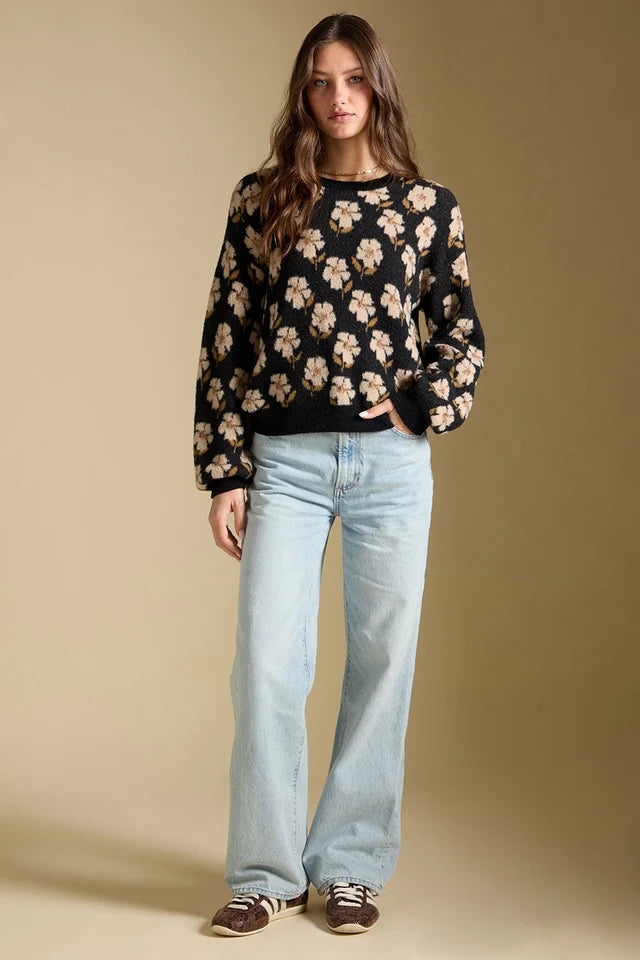 A woman modeling a black long-sleeve crewneck sweater with a beige and white floral pattern, paired with light-wash wide-leg jeans and brown sneakers, against a solid beige studio background.
