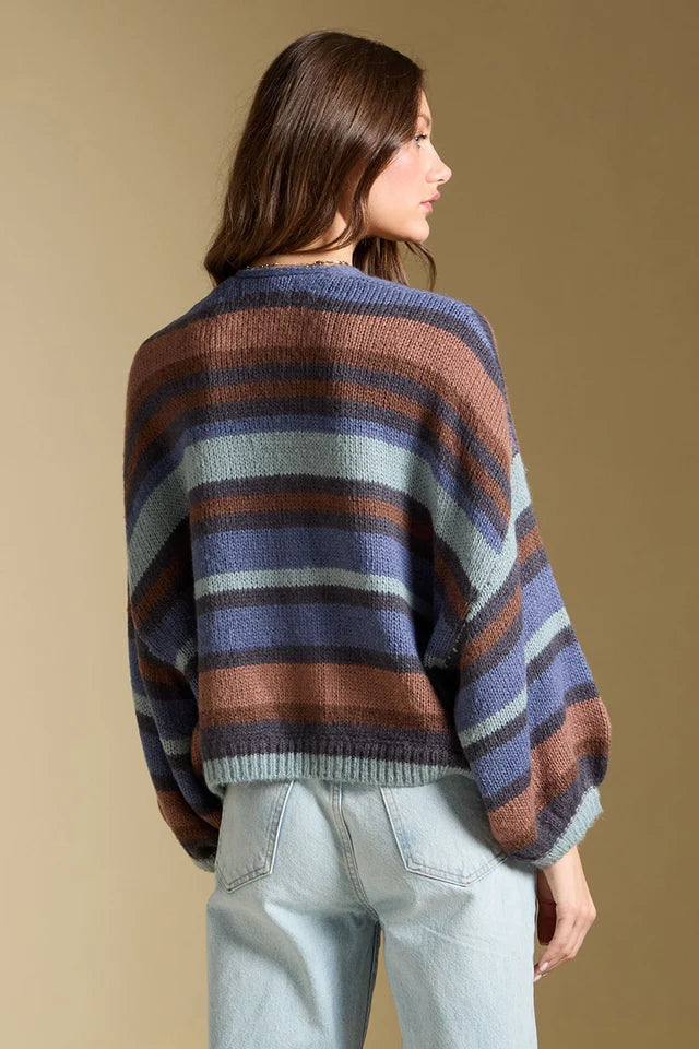 A woman shown from the back wearing a loose, oversized striped knit sweater with horizontal bands of blue, brown, beige, and dark navy, paired with light-wash jeans against a plain beige studio background.