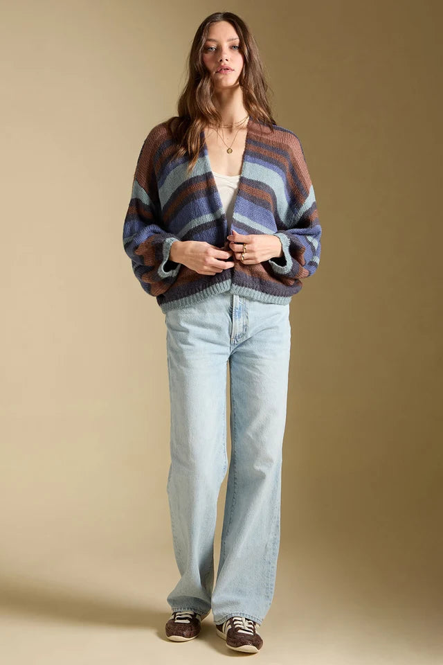A woman stands against a warm beige studio background wearing a striped knit cardigan in muted blue, brown, and rust tones, light-wash wide-leg jeans, and brown sneakers.