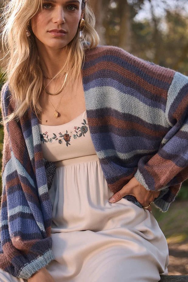 Woman wearing a striped cardigan over a white dress with floral embroidery, sitting outdoors.
