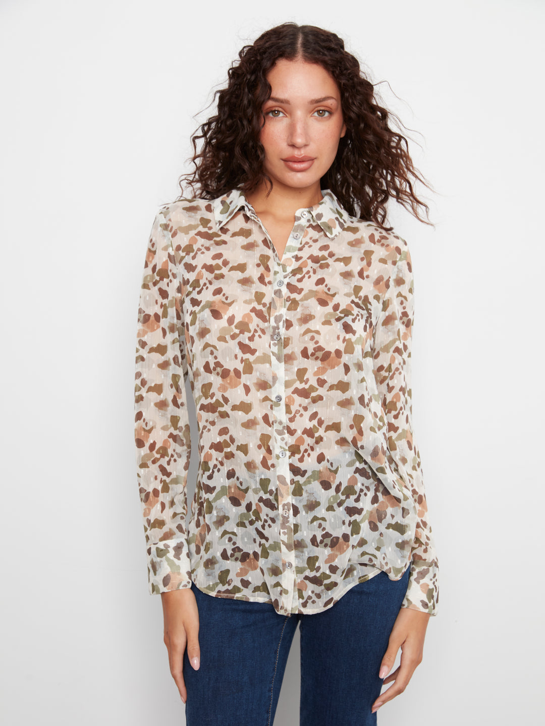 A woman with curly brown hair wearing a long-sleeve button-up blouse with a beige, brown, and gray leopard print, paired with dark blue jeans, against a plain white studio background.
