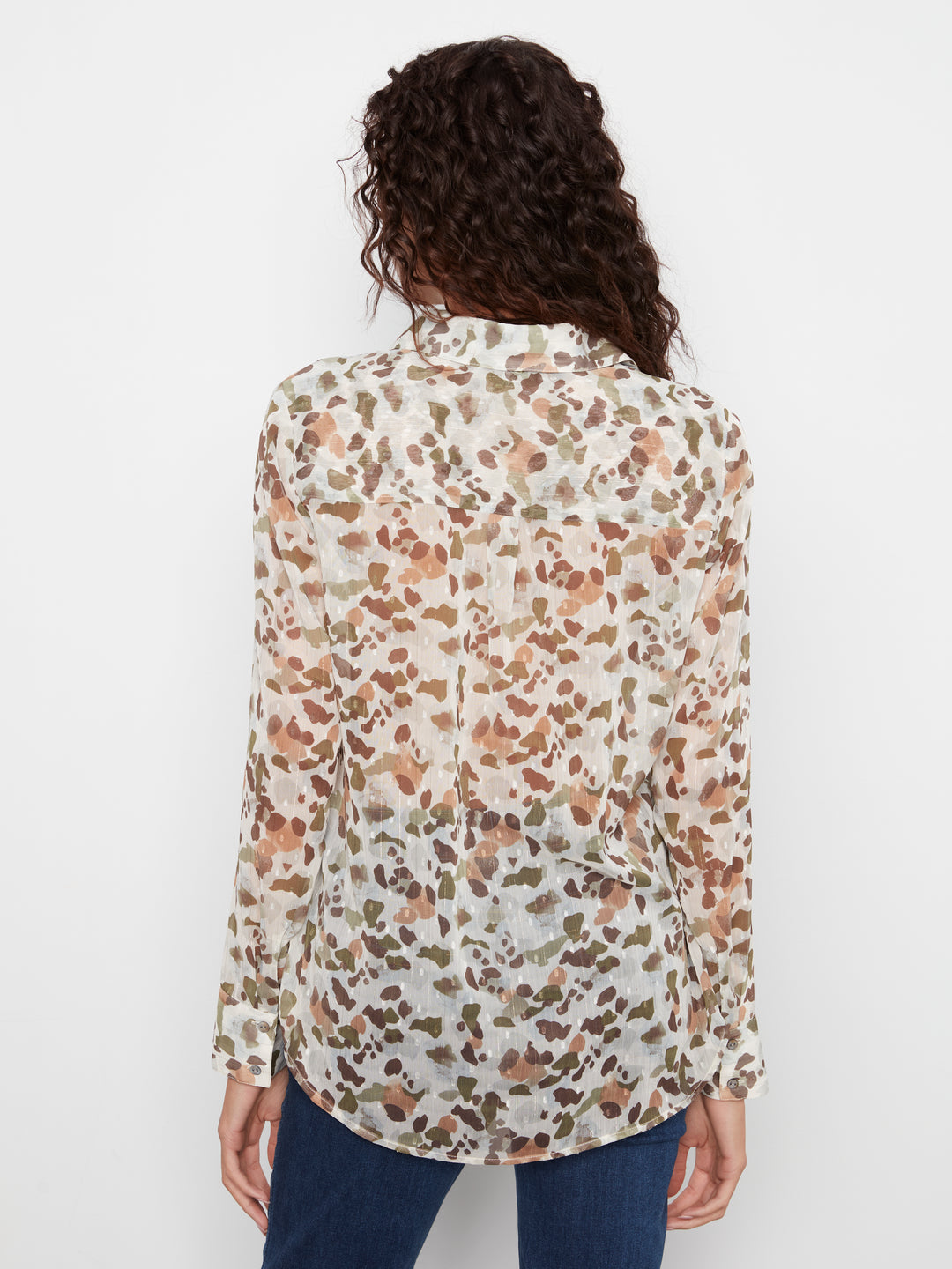 A woman viewed from the back wearing a long-sleeve button-up blouse with an abstract watercolor-style print in brown, beige, and muted green on an off-white base, paired with dark blue jeans against a plain white background.