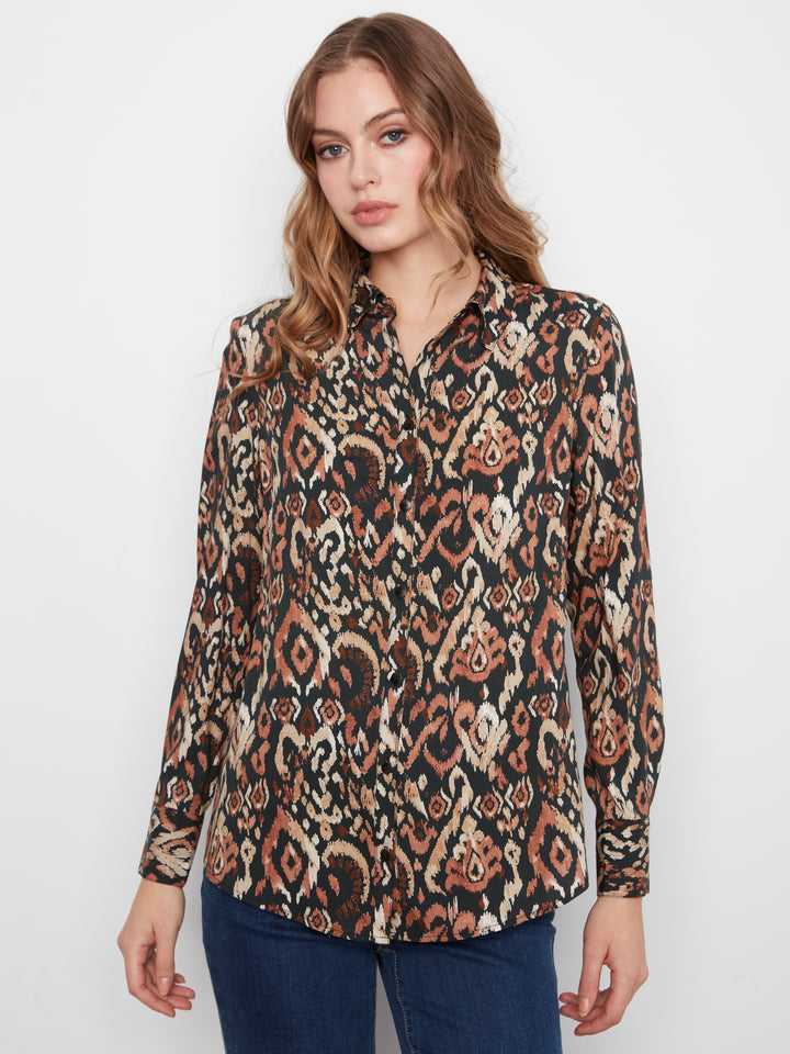 A woman wearing a long-sleeve button-up shirt with an intricate brown, beige, and black ikat pattern, paired with dark blue jeans, photographed against a plain white studio background.