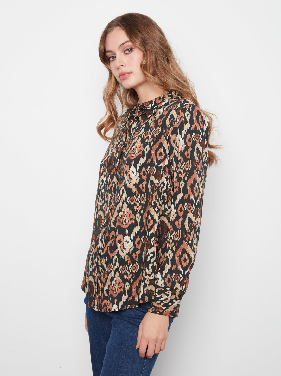 A woman wearing a long-sleeve button-up blouse with a brown, beige, and black geometric pattern, paired with dark blue jeans, photographed against a plain white studio background.