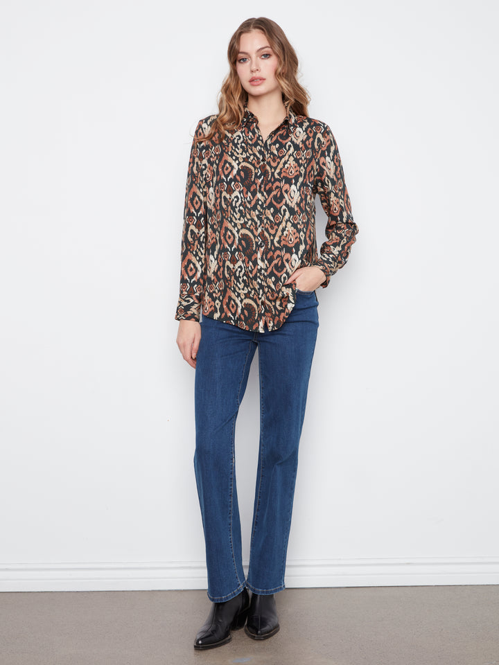A woman modeling a long-sleeve button-up shirt with a brown, beige, and black abstract pattern, paired with blue jeans and black boots, standing against a white wall.