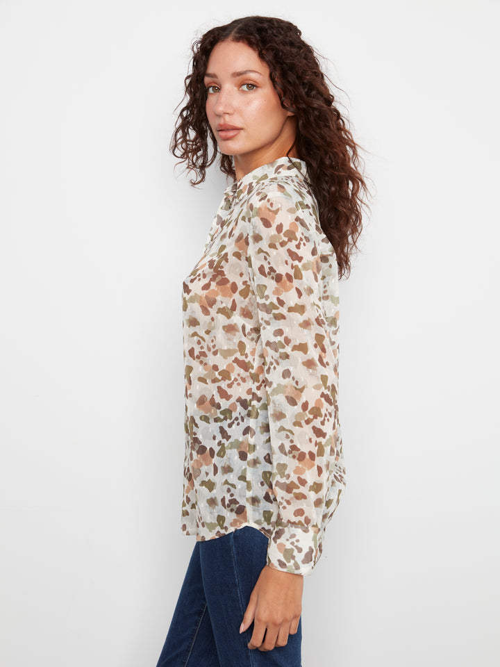A woman wearing a long-sleeve blouse with a brown and beige abstract print on a white background, paired with dark blue jeans, photographed against a light gray studio background.