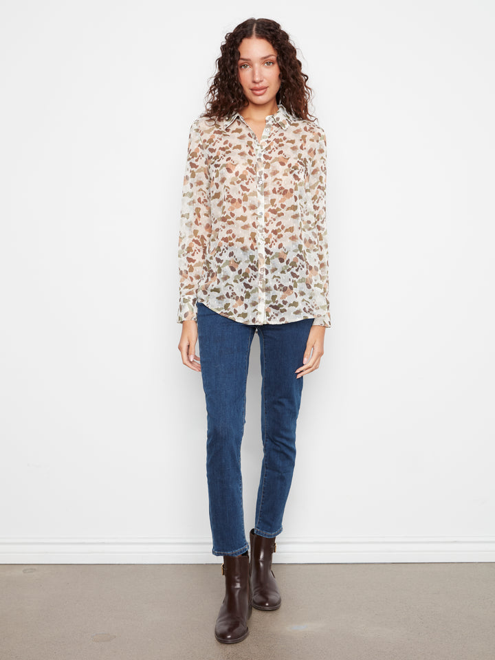 A woman stands against a white wall wearing a long-sleeve button-up blouse with a brown and beige floral print, blue jeans, and dark brown ankle boots.