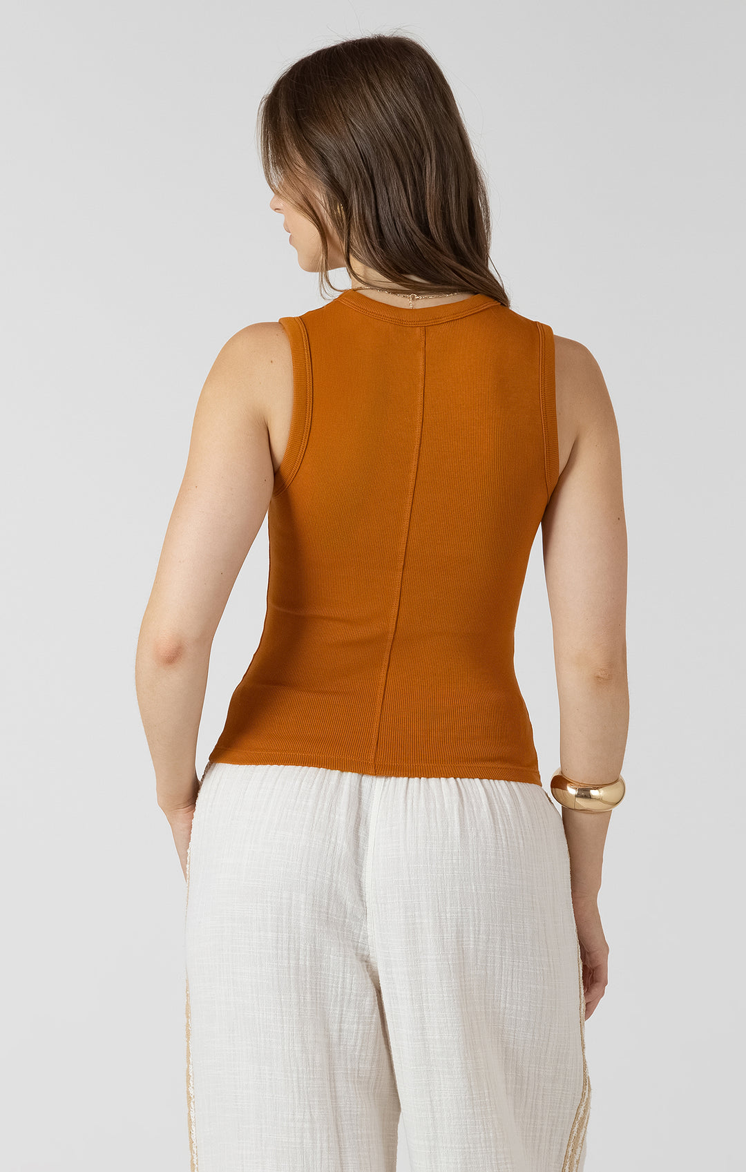 Woman wearing a mustard yellow sleeveless top and white pants on a light gray background