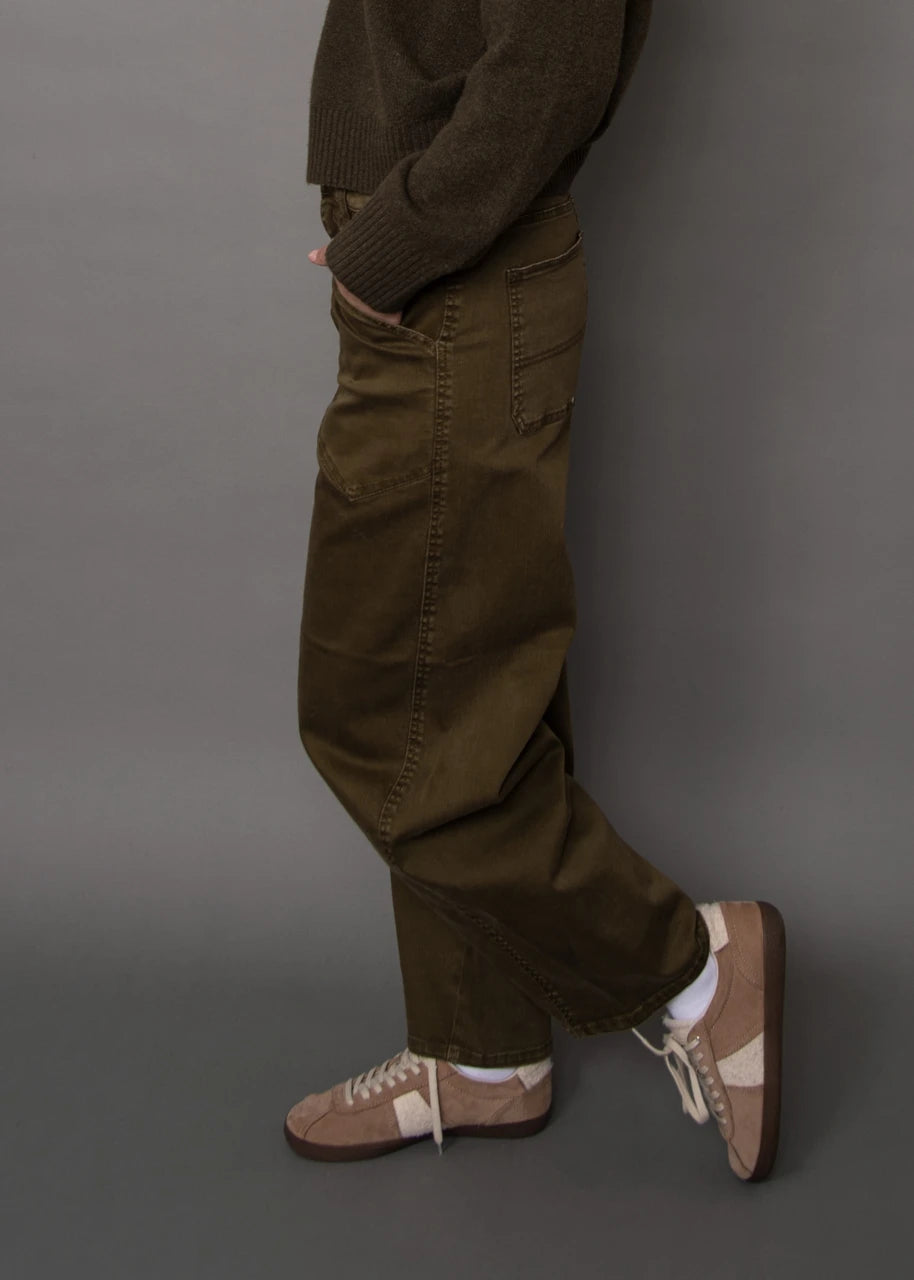 A person shown from the waist down wearing loose olive-brown corduroy pants, a dark brown knit sweater, white socks, and beige-and-brown sneakers against a plain gray studio background.