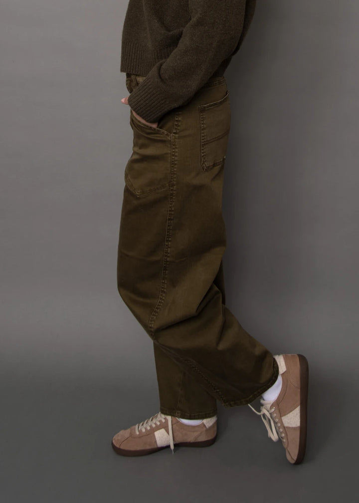 A person shown from the waist down wearing loose olive-brown corduroy pants, a dark brown knit sweater, white socks, and beige-and-brown sneakers against a plain gray studio background.