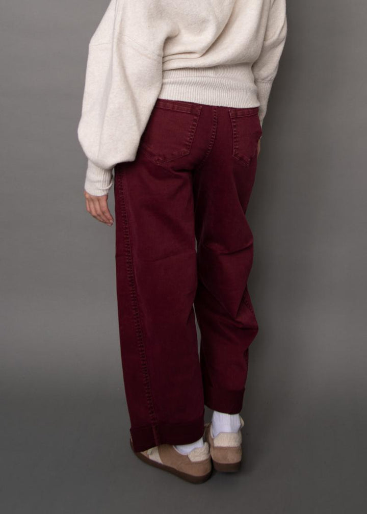 A person shown from the back wearing loose burgundy corduroy pants, a cream knit sweater, white socks, and tan slip-on shoes against a plain gray studio background.