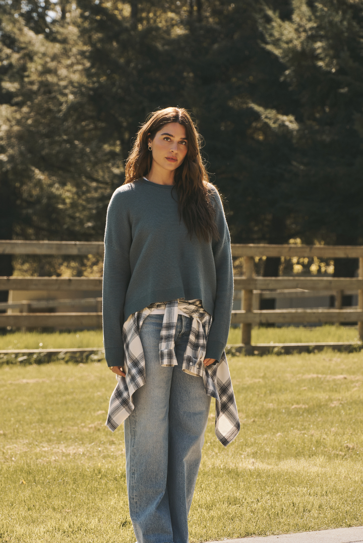 A woman standing outdoors in a grassy field with a wooden fence and trees in the background, wearing a muted blue-gray long-sleeve knit sweater, light-wash blue jeans, and a black-and-white plaid flannel shirt tied around her waist.