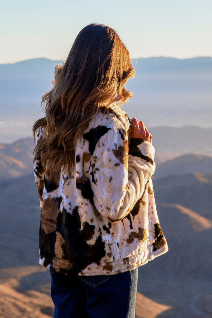 A woman with long brown hair stands on a mountain overlook, seen from behind, wearing a cream, black, and brown cow-print fleece jacket and blue jeans, looking out over a vast desert landscape under a clear sky.