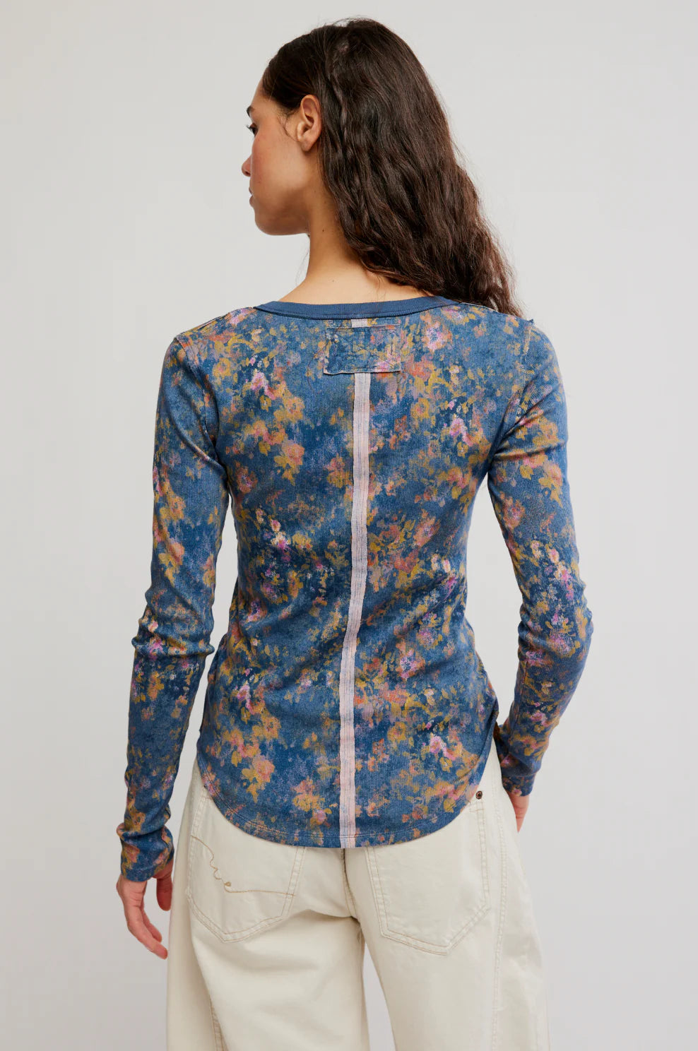 A woman shown from the back wearing a long-sleeve floral top with a blue base and multicolored floral print, paired with light beige pants, against a plain light gray studio background.