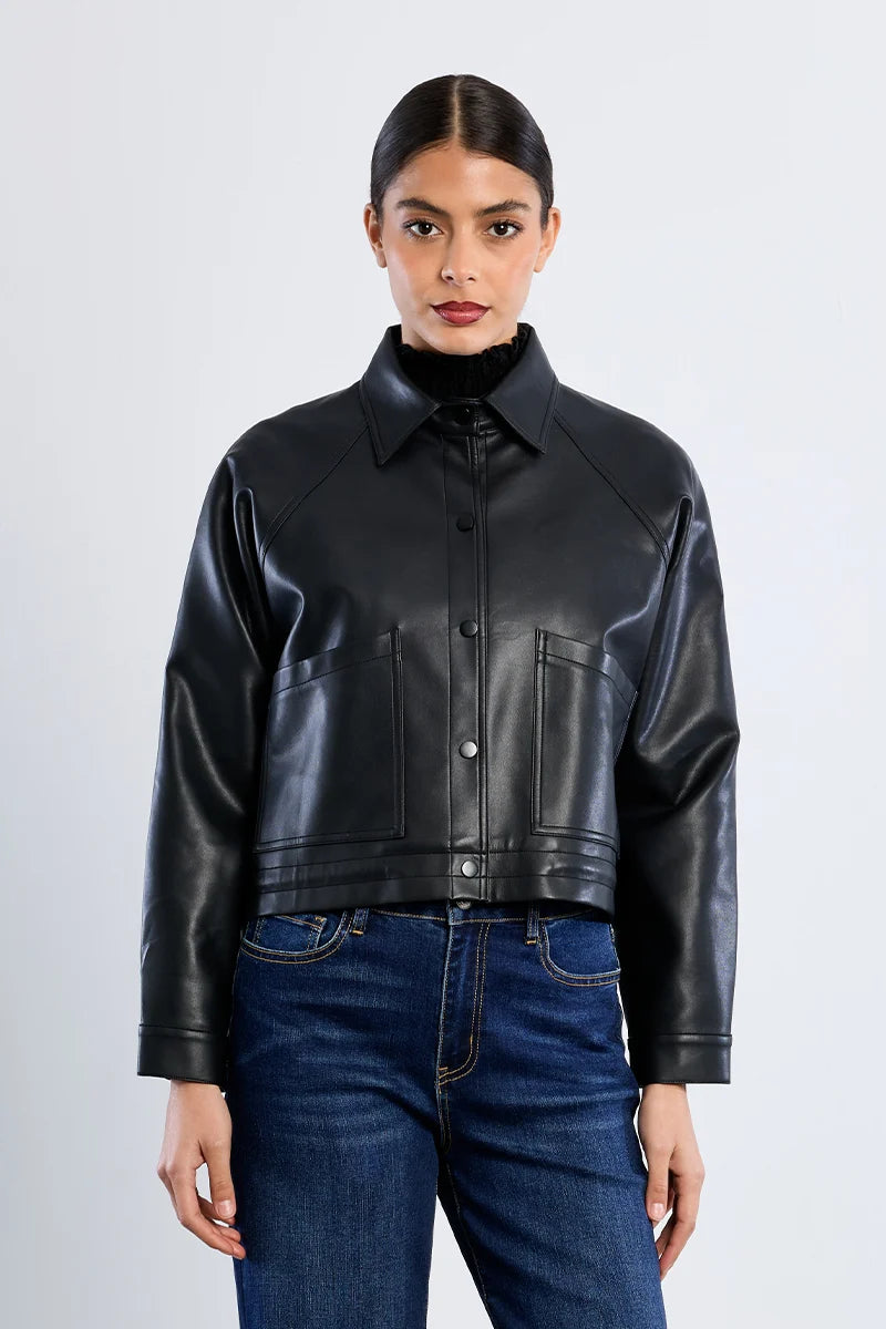 A model wearing a black leather cropped jacket with a high collar and front button closure, paired with blue jeans, against a light gray studio background.