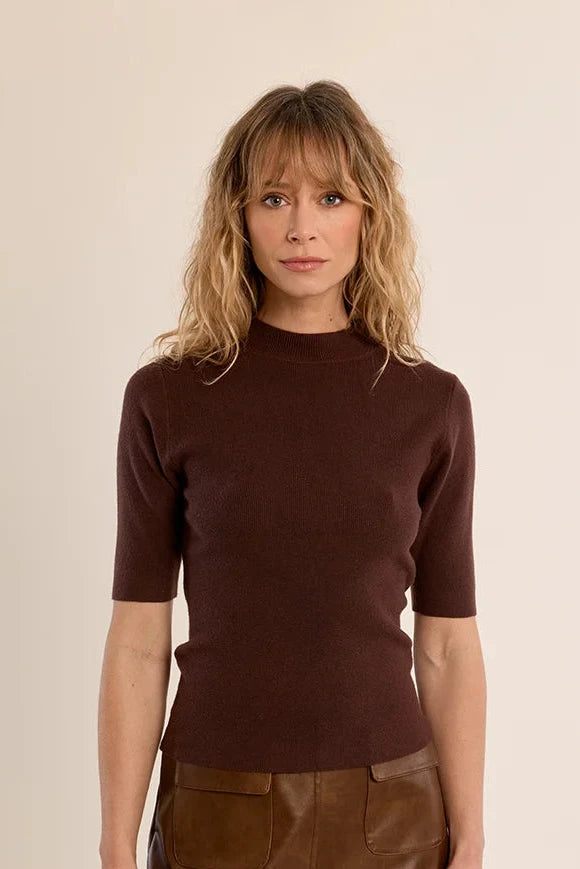 A woman modeling a dark brown short-sleeve knit top and a brown faux-leather mini skirt against a plain beige studio background.