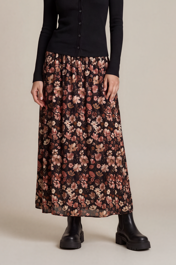 A woman stands against a plain beige studio background wearing a black long-sleeve button-front top and a long black skirt with a dense floral print in pink, beige, and muted orange tones, paired with black boots.