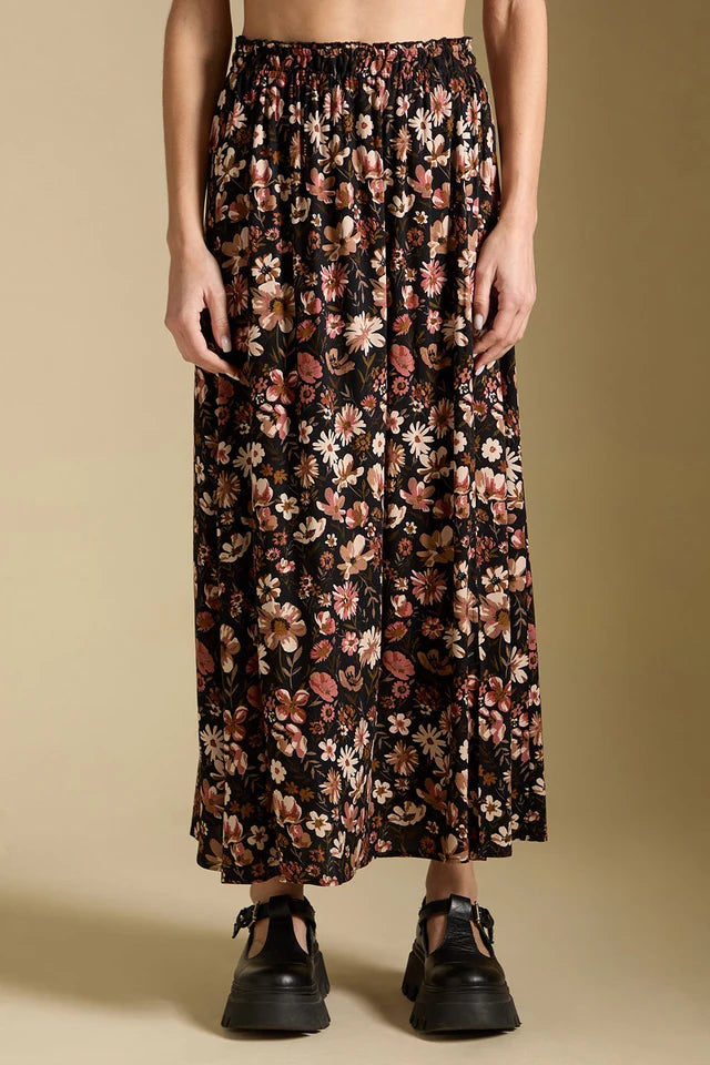 A model wearing a long black maxi skirt with a dense pink, beige, and white floral print, paired with black platform shoes, against a solid beige background.